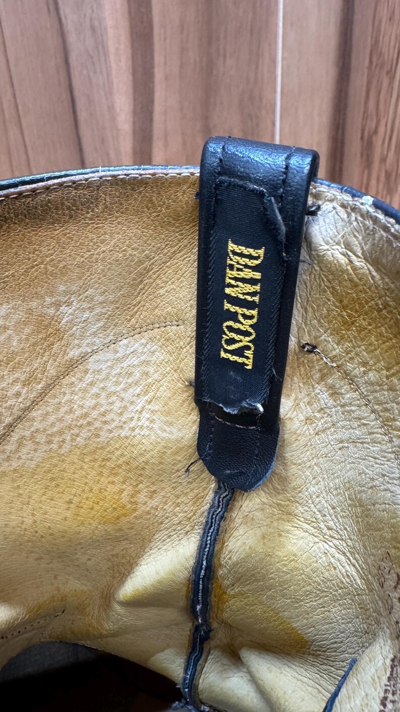 Op de afbeelding: Close-up van de binnenkant van een leren cowboylaars in bruin leer. Een zwarte treklus met de tekst "DAN POST" in gouden letters is zichtbaar. Het leer heeft een versleten uiterlijk.