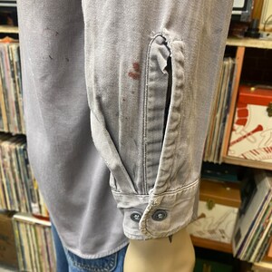 May include: A gray work shirt with long sleeves and two buttons on the cuff. The shirt has a few stains on it.