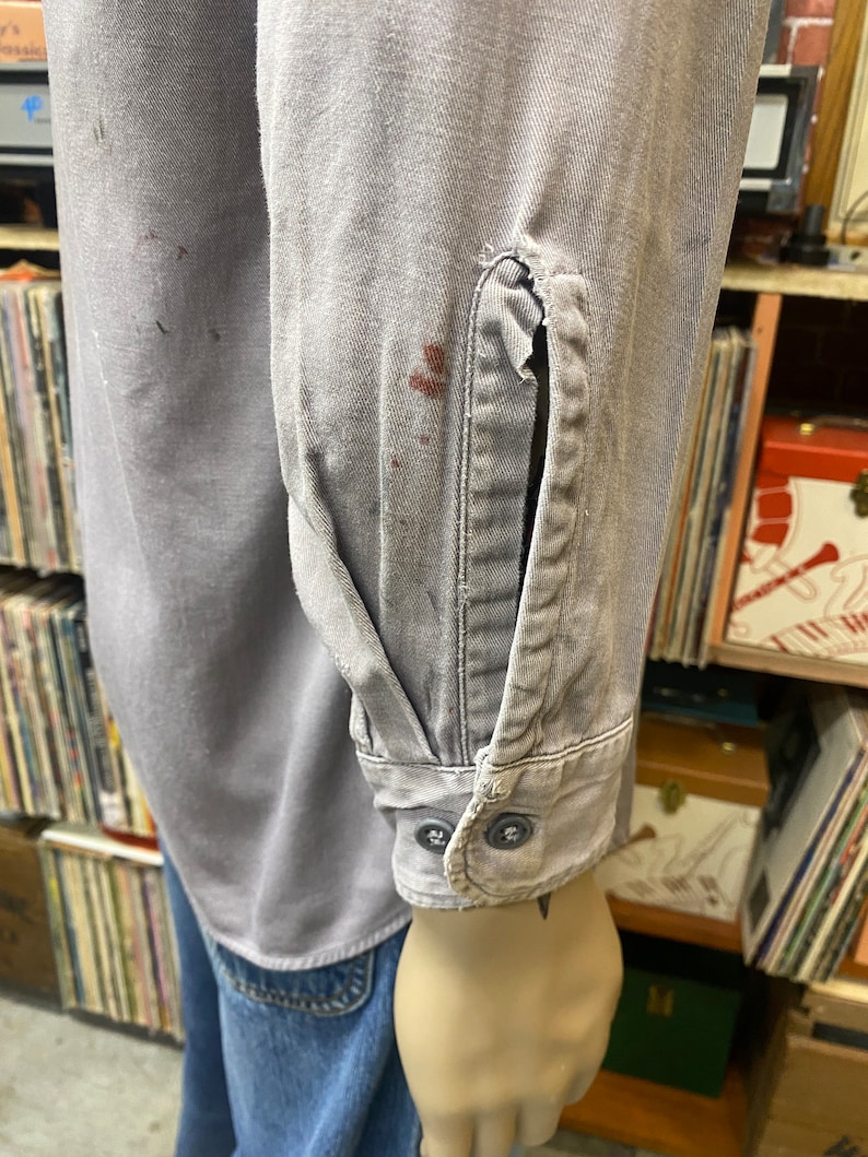 May include: A gray work shirt with long sleeves and two buttons on the cuff. The shirt has a few stains on it.