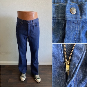 Puede incluir: Un par de pantalones vaqueros de mezclilla azul oscuro con cierre de cremallera y botón. Los pantalones tienen una abertura de pierna ancha y se muestran en un maniquí.