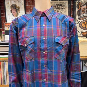 May include: A long-sleeved plaid shirt in shades of blue, red, and purple. The shirt features two chest pockets with button closures and a classic collar. The shirt is buttoned up the front with silver buttons.