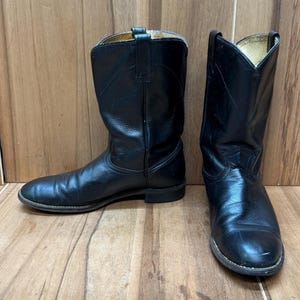 Op de afbeelding: Een paar zwarte leren cowboylaarzen. De laarzen hebben een klassiek ontwerp met een ronde neus, een middelhoge schacht en een gestapelde hak. Het leer heeft een lichte glans en de laarzen lijken versleten.