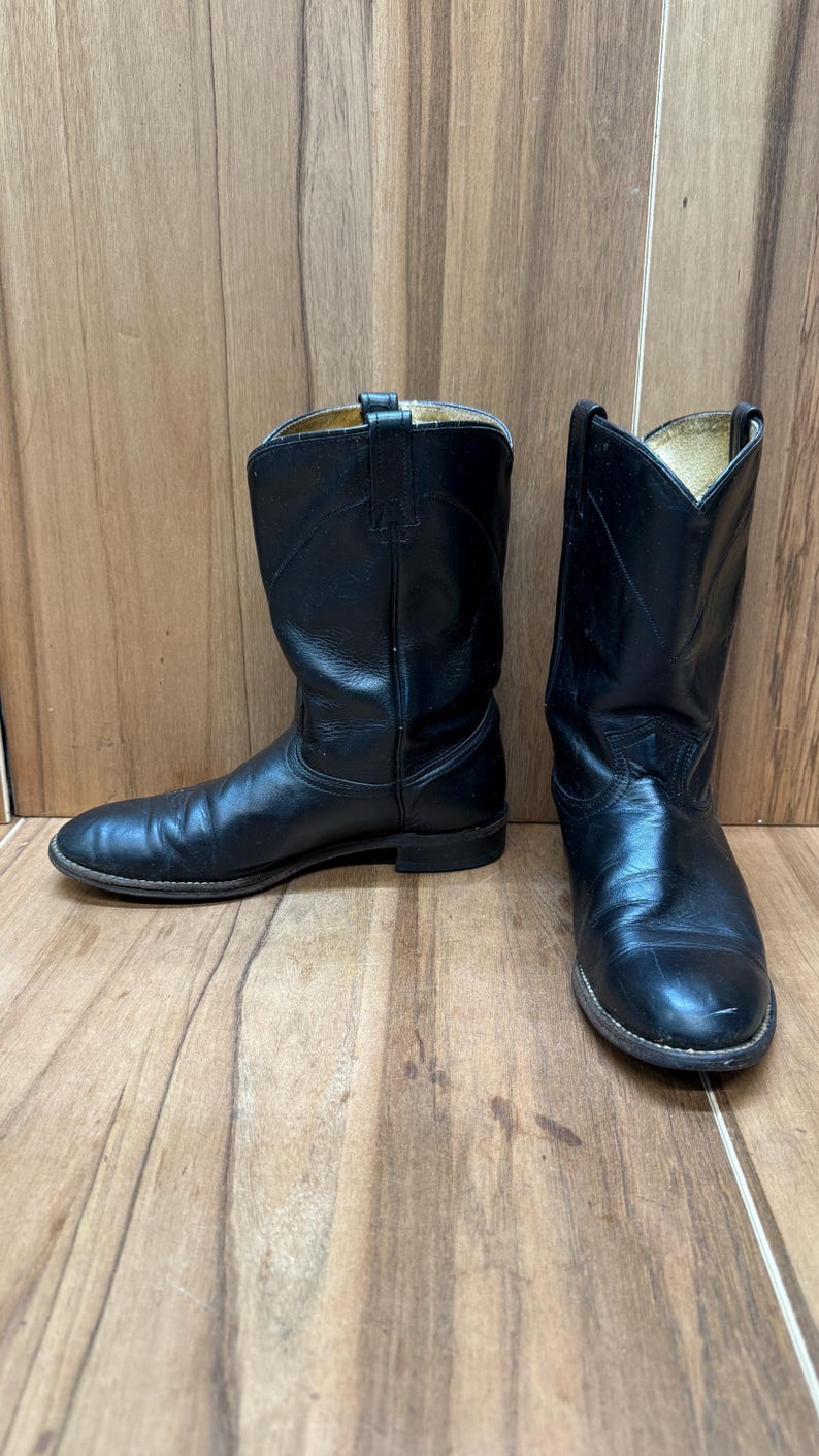 Op de afbeelding: Een paar zwarte leren cowboylaarzen. De laarzen hebben een klassiek ontwerp met een ronde neus, een middelhoge schacht en een gestapelde hak. Het leer heeft een lichte glans en de laarzen lijken versleten.