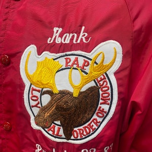 May include: Red jacket with a white embroidered name "Hank" and a white and brown embroidered moose patch with the text "Loyal Order of Moose" and "PAP". The jacket also has a white embroidered text "Prelate 82-83".