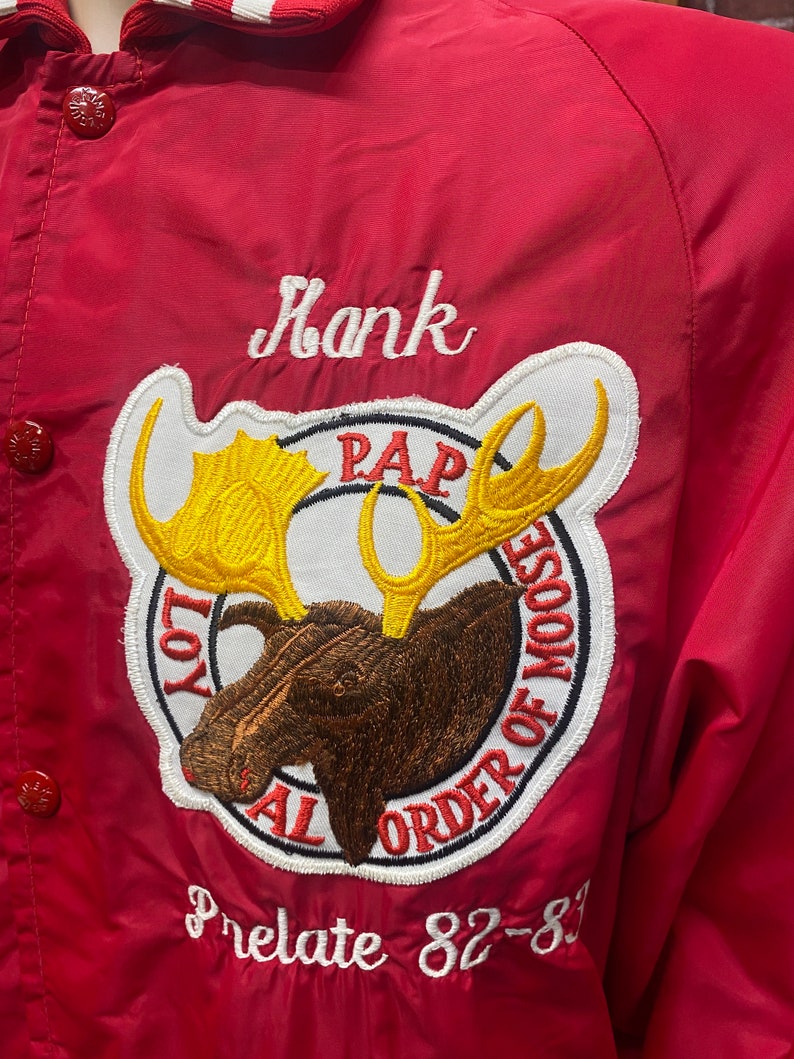 May include: Red jacket with a white embroidered name "Hank" and a white and brown embroidered moose patch with the text "Loyal Order of Moose" and "PAP". The jacket also has a white embroidered text "Prelate 82-83".