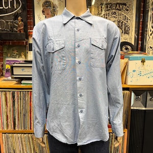 May include: A light blue long-sleeve button-down shirt featuring a classic collar, two chest pockets with buttoned flaps, and a central vertical row of matching blue buttons. The shirt has a structured fit and is displayed on a mannequin torso.