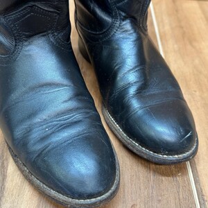 Op de afbeelding: Close-up van twee zwarte leren cowboylaarzen. De laarzen hebben een klassiek ontwerp met een puntige neus en een licht versleten uiterlijk. De zolen zijn lichtbruin.