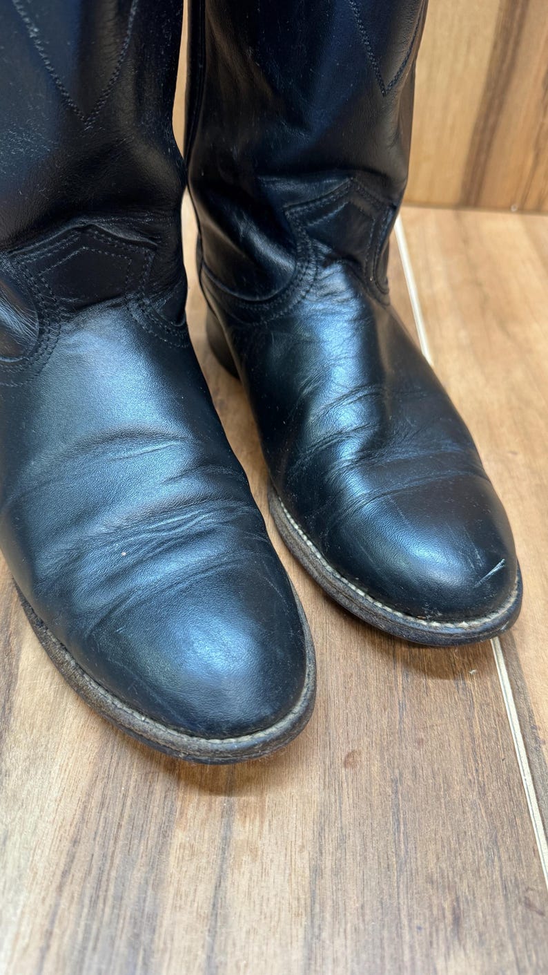 Op de afbeelding: Close-up van twee zwarte leren cowboylaarzen. De laarzen hebben een klassiek ontwerp met een puntige neus en een licht versleten uiterlijk. De zolen zijn lichtbruin.