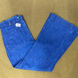 May include: Blue denim pants with a wide leg design. The pants have a button and zipper closure, with a back pocket and a small tag with the number "440". The pants are folded on a brown surface.