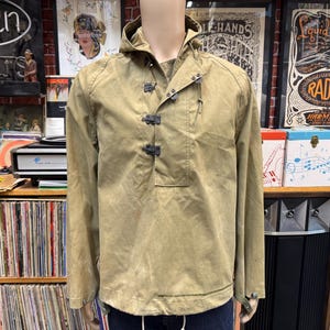 May include: A tan hooded pullover jacket featuring a high neckline with a drawstring, long sleeves, and a front placket secured by three dark metal hook-and-loop fasteners. The garment has a straight hemline and a structured, utilitarian design.