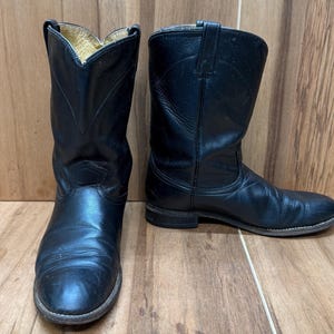Op de afbeelding: Een paar zwarte leren cowboylaarzen. De laarzen hebben een klassiek ontwerp met een puntige neus en een middelhoge kuit. Het leer lijkt versleten, met zichtbare kreukels en slijtplekken. De laarzen staan tegen een houten achtergrond.