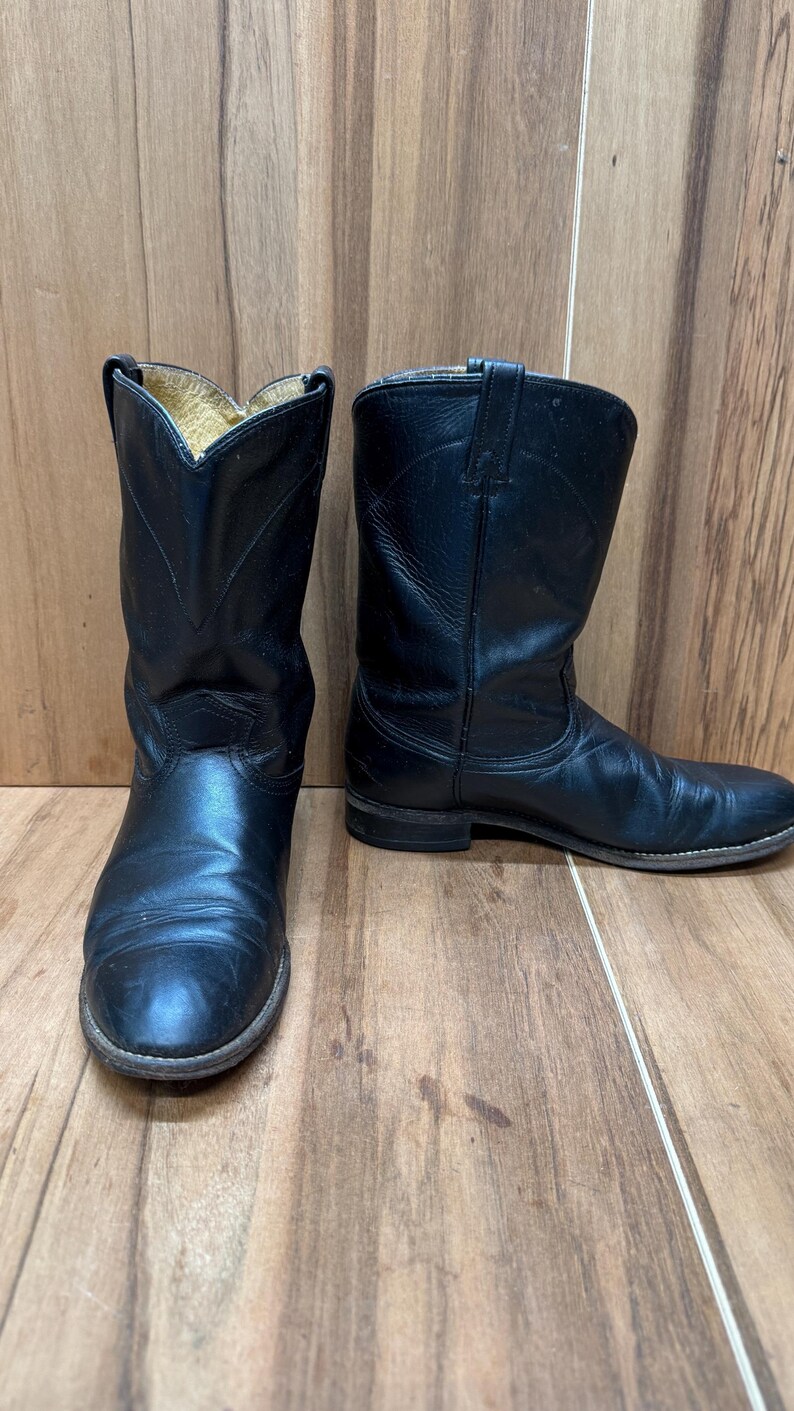 Op de afbeelding: Een paar zwarte leren cowboylaarzen. De laarzen hebben een klassiek ontwerp met een puntige neus en een middelhoge kuit. Het leer lijkt versleten, met zichtbare kreukels en slijtplekken. De laarzen staan tegen een houten achtergrond.