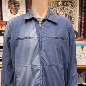 May include: A blue long-sleeve zip-up jacket with a pointed collar, two horizontal zippered chest pockets, and a visible vertical seam running down the center. The jacket is worn over a white and black horizontal striped shirt underneath.