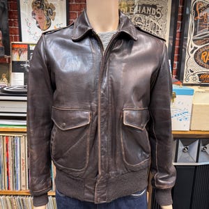 May include: A dark brown leather bomber jacket featuring a classic collar, a full-length front zipper, and two large flap pockets on the front. The jacket includes ribbed cuffs and a ribbed hem, with shoulder epaulets and a smooth leather finish.