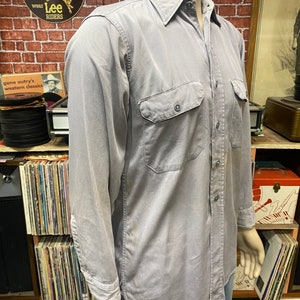 May include: A light gray long-sleeved work shirt with a button-down collar, two chest pockets, and a single button cuff. The shirt is made of a durable, cotton fabric.