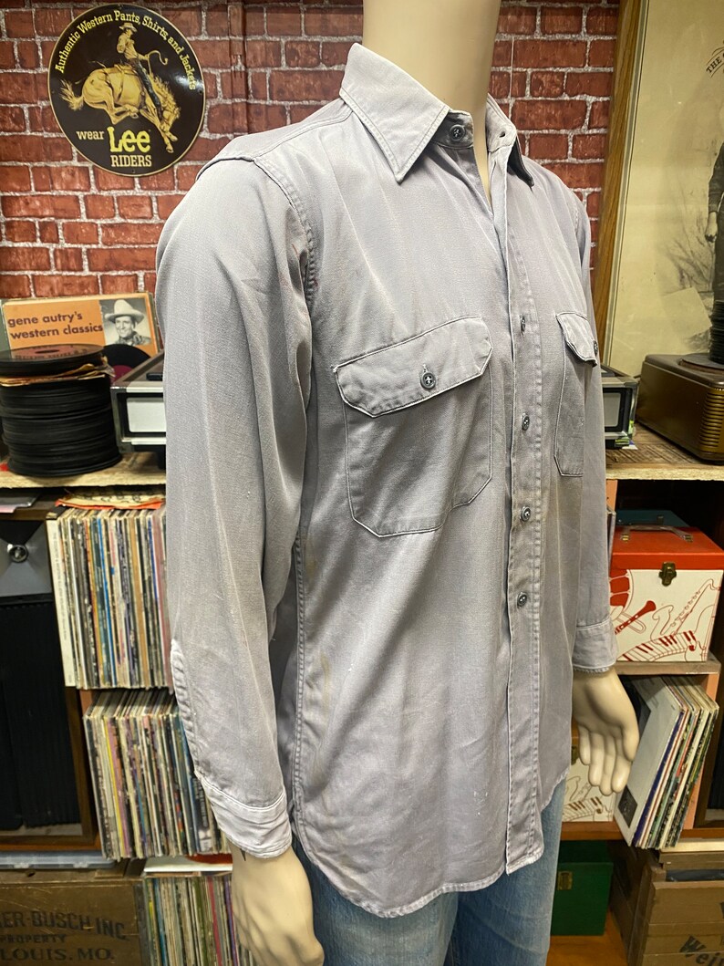 May include: A light gray long-sleeved work shirt with a button-down collar, two chest pockets, and a single button cuff. The shirt is made of a durable, cotton fabric.
