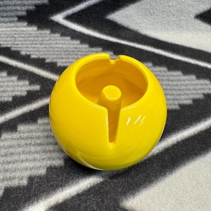 May include: A bright yellow ceramic ashtray with a round shape and a small hole in the center. The ashtray is sitting on a black and white patterned surface.