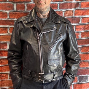 May include: A black leather motorcycle jacket featuring a wide notched collar, silver-toned metal zippers, a diagonal chest pocket, and a buckled waist belt. The jacket includes shoulder epaulets and long sleeves with zippered cuffs, providing a structured fit.