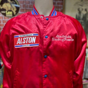 May include: Red satin bomber jacket with blue and white striped trim. The jacket has a patch on the left chest that reads "Factory Trained Alston Dealer" and embroidered text on the right chest that reads "Stan Goldstein Director of Promotion."