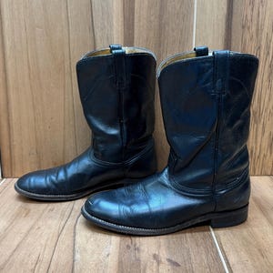 Op de afbeelding: Een paar zwarte leren cowboylaarzen. De laarzen hebben een klassiek ontwerp met een ronde neus en een middelhoge schacht. Het leer vertoont tekenen van slijtage, wat de schoenen karakter geeft. De laarzen worden op een houten oppervlak getoond.