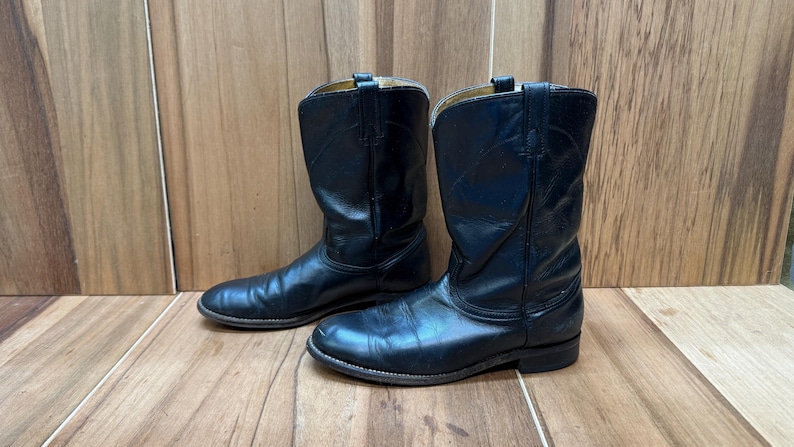 Op de afbeelding: Een paar zwarte leren cowboylaarzen. De laarzen hebben een klassiek ontwerp met een ronde neus en een middelhoge schacht. Het leer vertoont tekenen van slijtage, wat de schoenen karakter geeft. De laarzen worden op een houten oppervlak getoond.