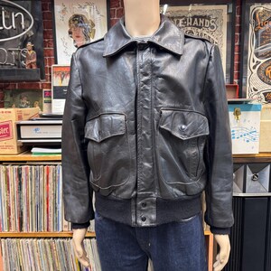 May include: A black leather bomber jacket with two front pockets and a ribbed hem and cuffs. The jacket has a collared neckline and is displayed on a mannequin. The jacket is a classic style.