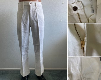 May include: White trousers featuring a high-waisted design, a metal button closure, and a brass-toned zipper fly. The pants include belt loops and show visible dark smudges on the lower leg area. The interior waistband displays a faded tag.