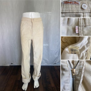 May include: A pair of off-white corduroy pants with a zipper fly and button closure. The pants have a straight leg and are cuffed at the bottom. The pants have a Levi's label on the back pocket and a "Worn One" label on the waistband.