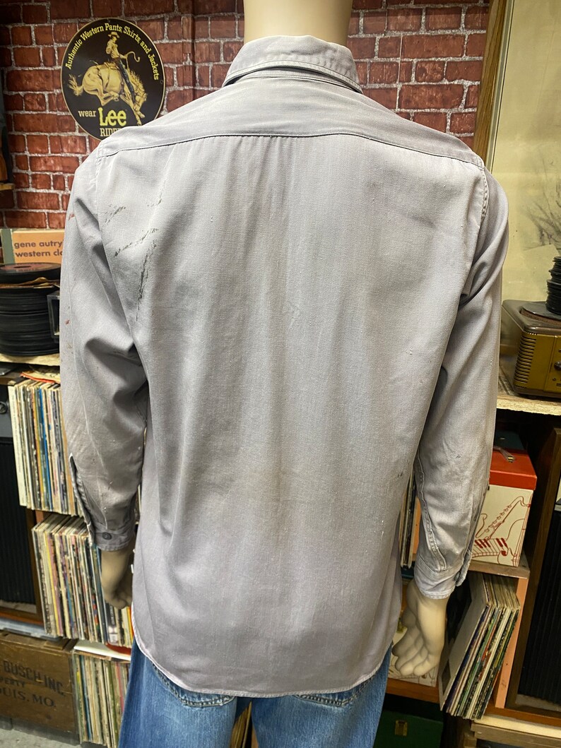 May include: A gray long-sleeved work shirt with a button-down collar. The shirt has a faded appearance and is slightly wrinkled.