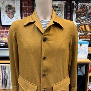 May include: A mustard yellow long-sleeved jacket with a button-up front and two large pockets. The jacket has a collar and is made of a soft, textured fabric.