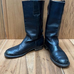 May include: A pair of black leather boots. The boots are knee-high with a rounded toe and a low heel. The leather appears worn, with visible creases and scuffs. The boots are standing on a wooden surface.