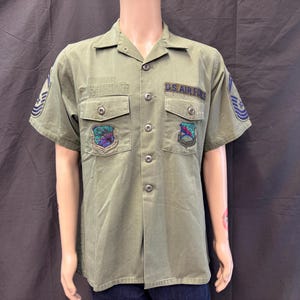 May include: An olive green short-sleeve military shirt featuring a button-down front, two chest pockets, and embroidered patches. The shirt displays the text U.S. AIR FORCE above the right pocket, with additional insignia on the sleeves and chest.