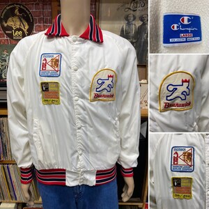 May include: White satin bomber jacket with red and blue trim. The jacket has several embroidered patches, including a Brunswick bowling logo, a Champion logo, and a Member of the American Junior Bowling Congress logo. The jacket also has a red and blue striped collar.