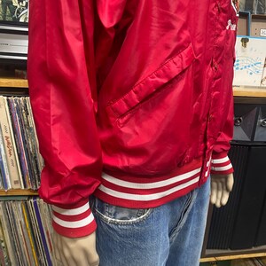 May include: A red satin baseball jacket with white and red striped cuffs and waistband. The jacket has a button closure and a pocket on the left side. The jacket has the word "Princeton" embroidered on the right side.