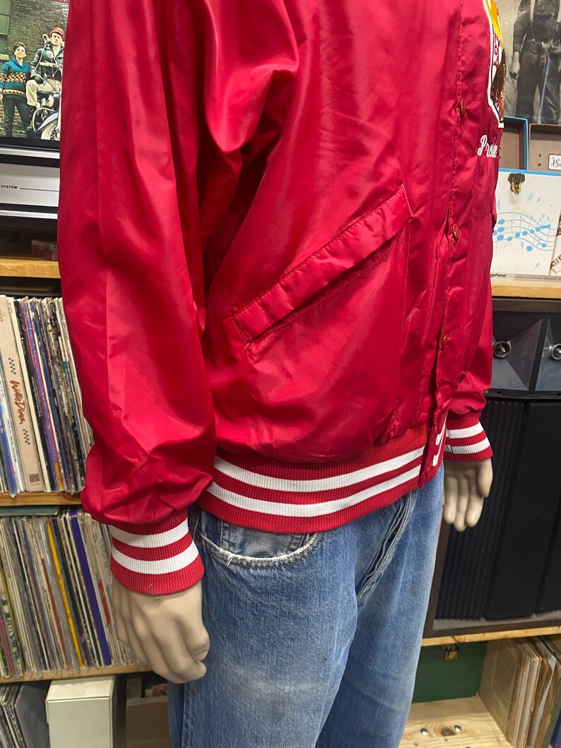 May include: A red satin baseball jacket with white and red striped cuffs and waistband. The jacket has a button closure and a pocket on the left side. The jacket has the word "Princeton" embroidered on the right side.