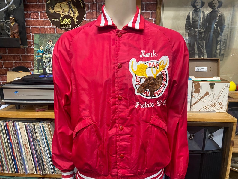 May include: Red satin bomber jacket with white and red striped trim. The jacket features an embroidered moose with the text "Loyal Order of Moose" and "Prelate 82-83".