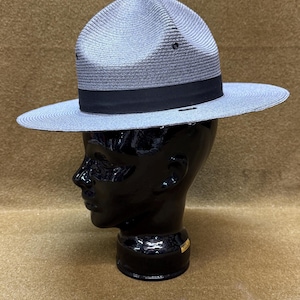 May include: A grey straw campaign hat with a high, pinched crown and a wide, flat brim. The hat features a black grosgrain ribbon band encircling the base of the crown and small metal ventilation eyelets on the sides of the crown structure.