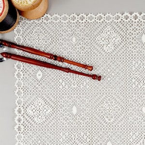 Może przedstawiać: Biała koronka szydełkowa z delikatnym wzorem. Na koronce leżą dwa drewniane nawijacze.