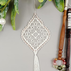 Può includere: Ornamento in pizzo all'uncinetto bianco con un fiocco che pende da un soffitto bianco. L'ornamento ha la forma di un diamante con un motivo floreale.