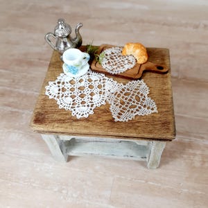 May include: A miniature wooden table with a distressed white base and brown top, featuring a silver teapot, a small teacup, a wooden serving board with a croissant, and three intricate white crocheted doilies arranged on the surface.