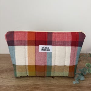 May include: A rectangular quilted zipped pouch featuring a multi-coloured plaid pattern in shades of red, cream, blue, green, and mustard yellow. A white fabric label centred on the front displays the black text these hands in lowercase.