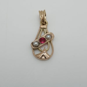 May include: A gold pendant with a teardrop shape and a delicate design. The pendant features three small pearls and a red gemstone.