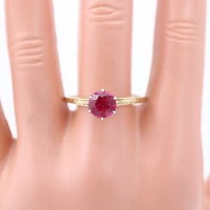May include: A gold ring featuring a large, round, red ruby gemstone set in a prong setting. The ring has a delicate, engraved band.