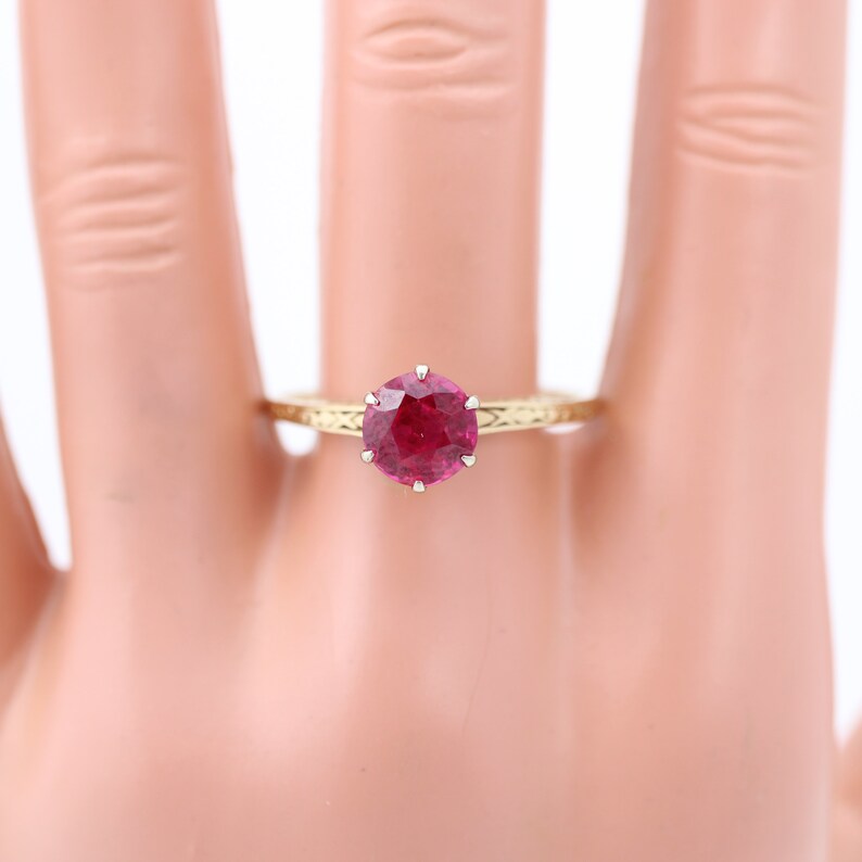 May include: A gold ring featuring a large, round, red ruby gemstone set in a prong setting. The ring has a delicate, engraved band.