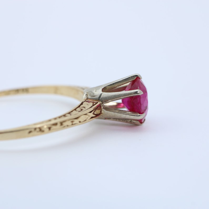 May include: A gold ring with a red ruby gemstone set in a prong setting. The band is decorated with an engraved floral design.
