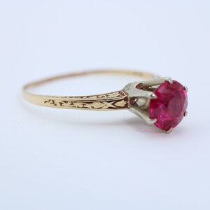 May include: A gold ring with a bright red ruby gemstone set in a white metal prong setting. The band is engraved with a delicate floral design.