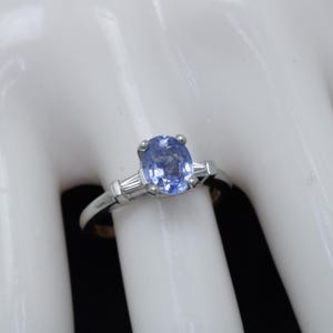 Puede incluir: Un anillo de plata con una piedra de zafiro ovalada azul claro y dos diamantes más pequeños de corte baguette a los lados.