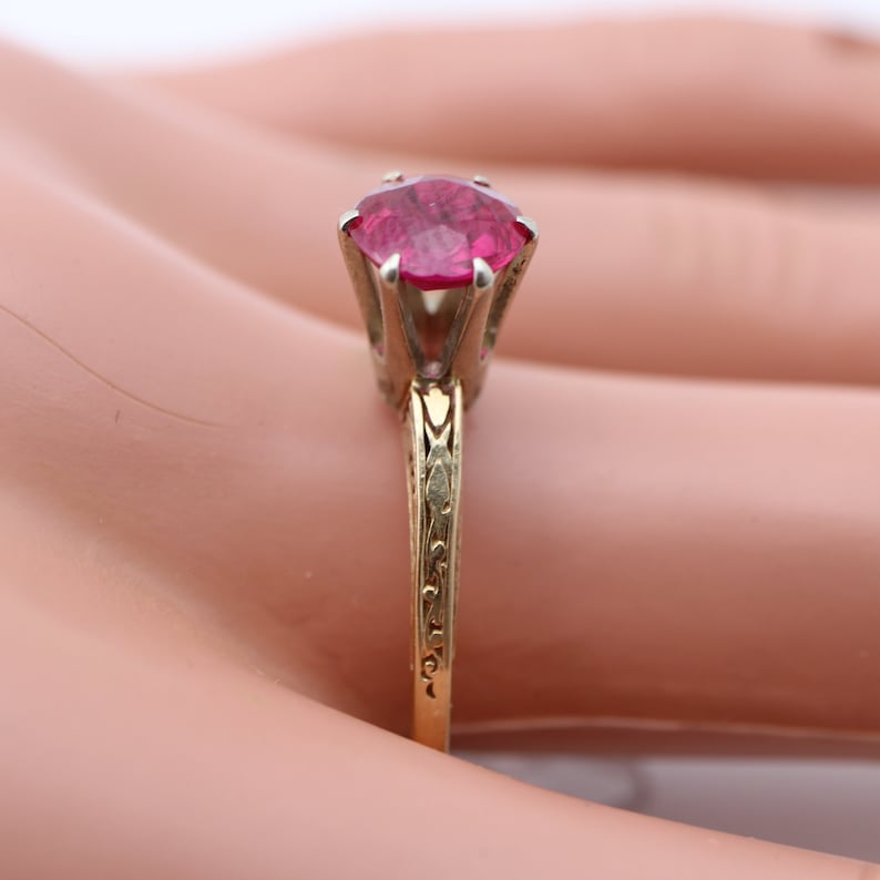 May include: A gold ring featuring a pink gemstone set in a prong setting. The band is adorned with a delicate floral design.