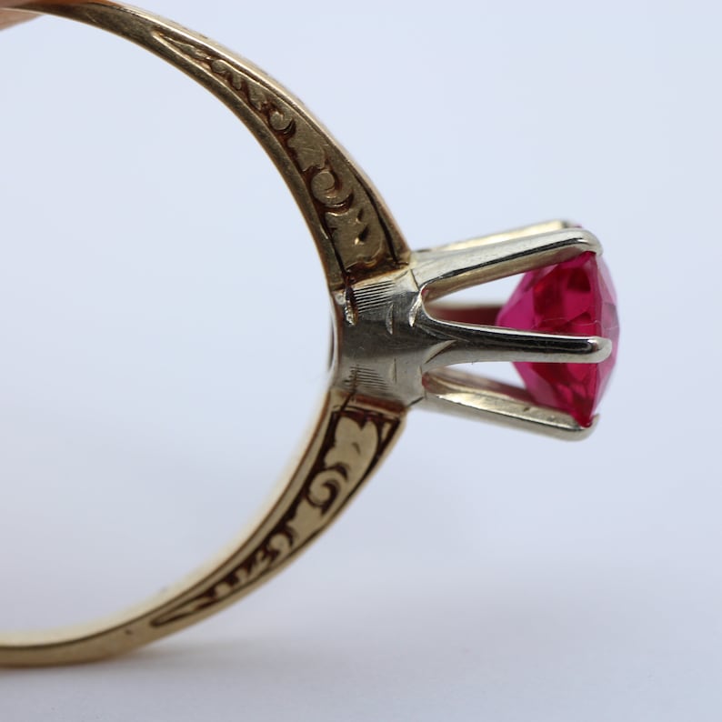 May include: A gold ring with an ornate design on the band and a red gemstone set in a prong setting.