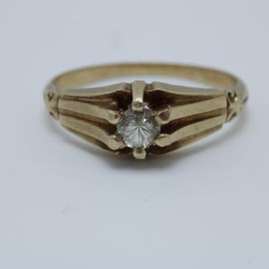 May include: A gold ring with a single round diamond set in the center. The band is decorated with three raised lines and a floral design on each side.
